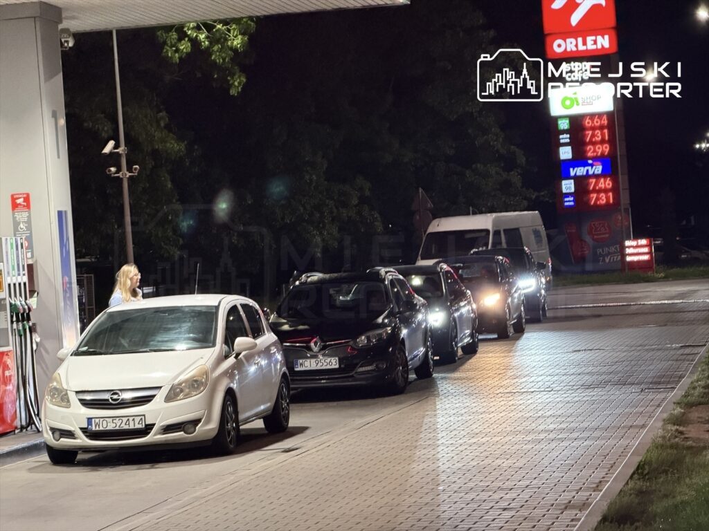 Kobieta z długimi włosami zbliża się do białego samochodu osobowego na stacji paliw, obok czeka rząd aut.