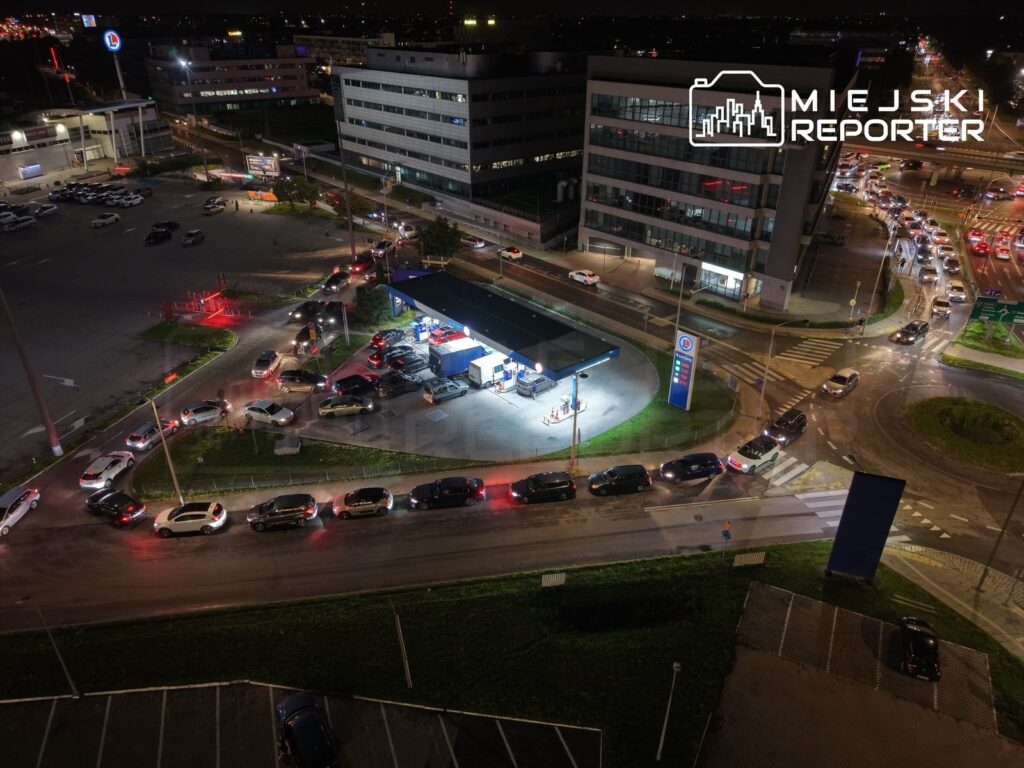 Samochody w długiej kolejce czekają na zatankowanie na stacji paliw w nocy, otoczone oświetlonymi budynkami.
