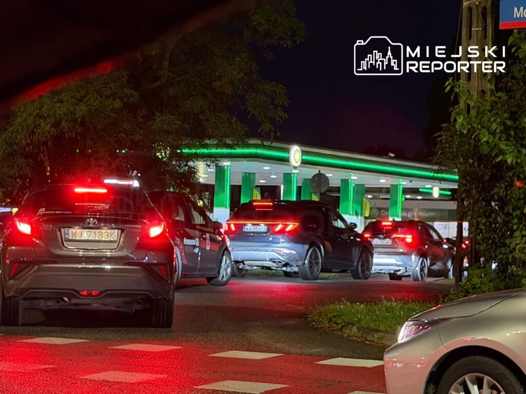 Samochody stoją w długiej kolejce przed stacją paliw, oświetloną zielonymi światłami, w nocy.