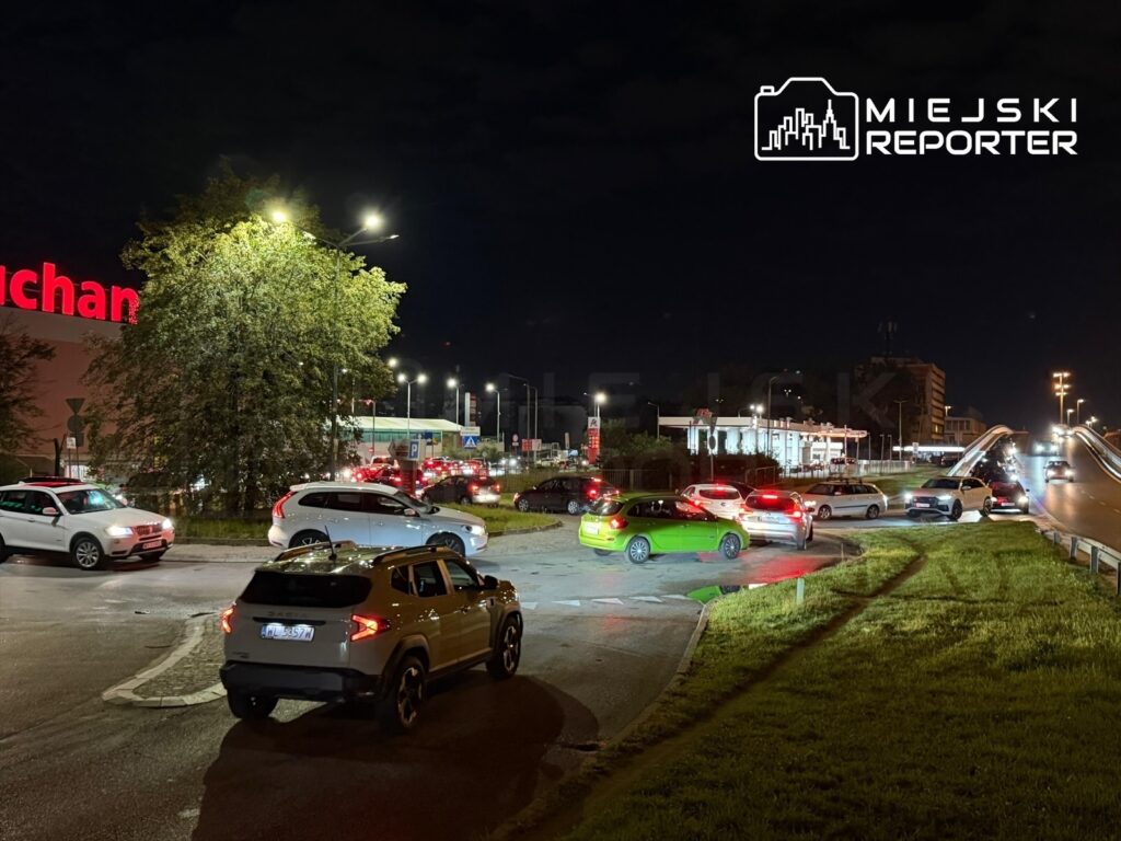 Samochody stoją w długim korku na rondzie w pobliżu stacji paliw, w tle widoczny jest jasny neon supermarketu.