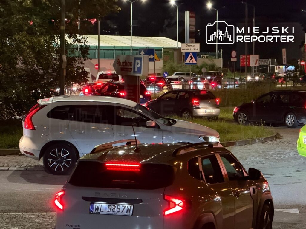 Ruchliwe skrzyżowanie z samochodami czekającymi w kolejce do stacji paliw, oświetlone nocnymi latarniami.