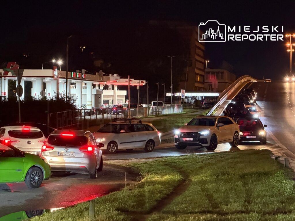 Ruch samochodowy przed stacją paliw w nocy, z pojazdami czekającymi w długiej kolejce na zatankowanie.