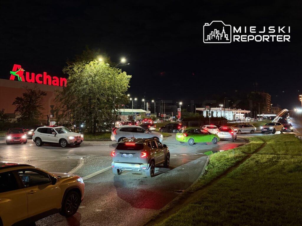 Ruchliwa ulica z samochodami czekającymi w korku, w tle wyraźnie widoczny neonowy napis "Auchan" oraz stacja paliw.