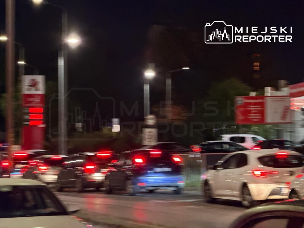 Ruchliwa stacja paliw w nocy, z długą kolejką samochodów oczekujących na tankowanie.
