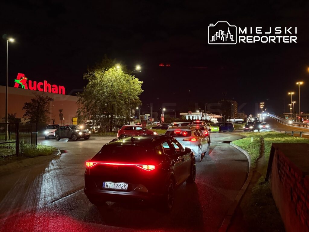 Ruch samochodowy przed hipermarketem Auchan w nocy, z widocznymi światłami i kolejką pojazdów.