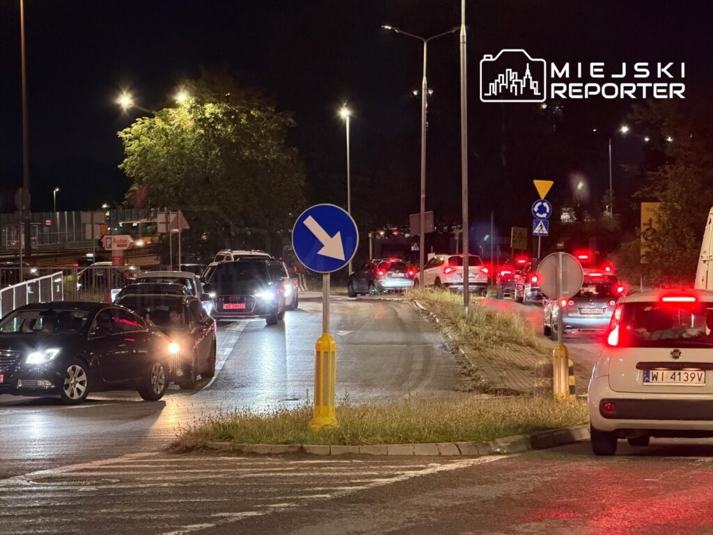 Ruch samochodowy na skrzyżowaniu w nocy, z pojazdami oczekującymi na wjazd na stację paliw.