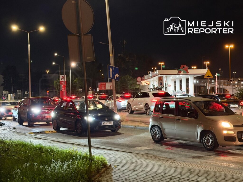 Ruch samochodowy w nocy przed stacją paliw, z kilkoma pojazdami oczekującymi w kolejce.