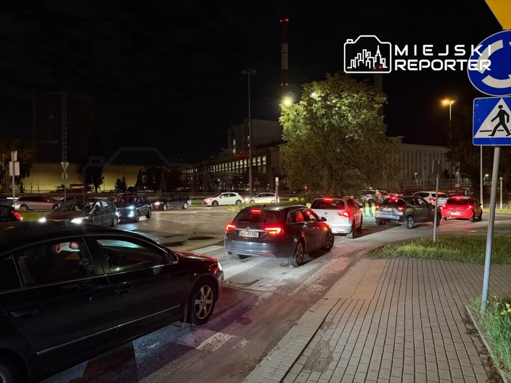 Ruchliwa ulica nocą, z długim korkiem samochodowym w kierunku stacji paliw, otoczona oświetlonymi budynkami.