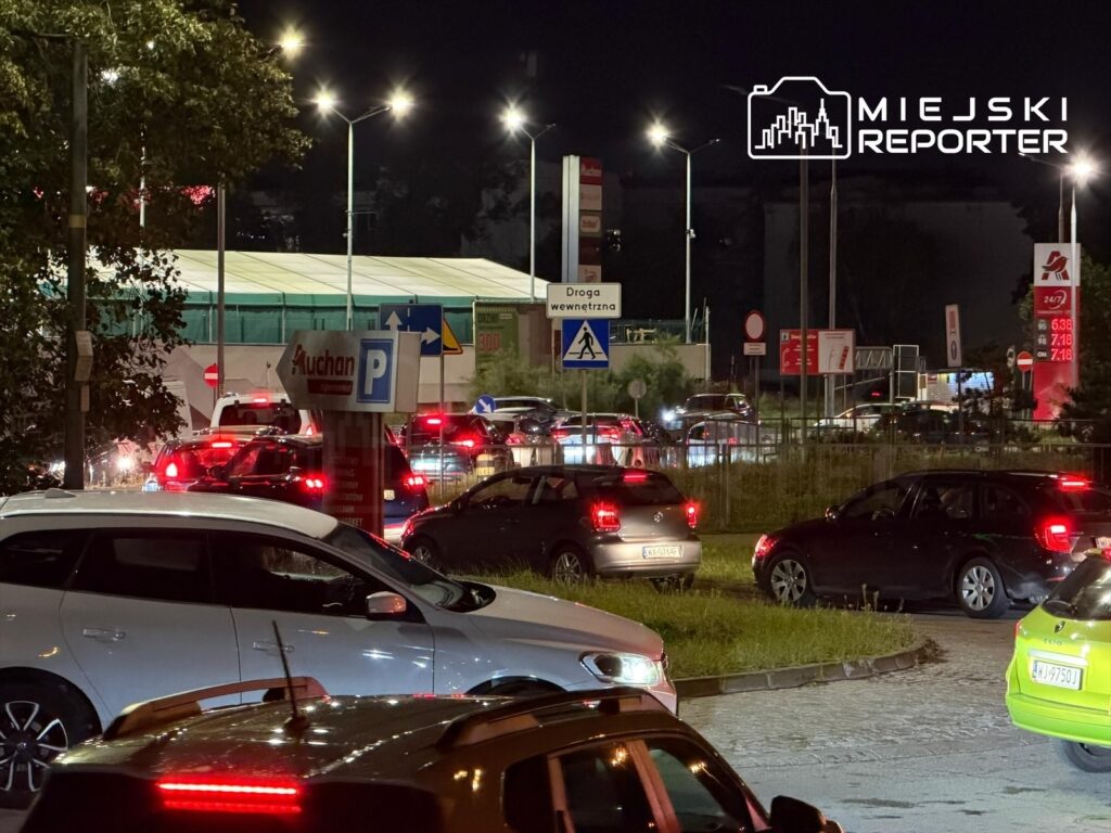 Ruchliwy parking przed stacją paliw w nocy, z samochodami czekającymi w kolejce na tankowanie. W tle widoczne znaki drogowe.