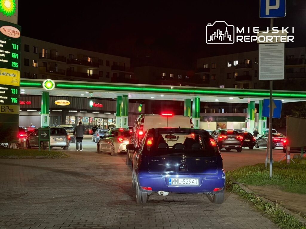 Kierowcy czekają w długiej kolejce do stacji paliw BP w nocy, z widoczną tablicą cen paliw.