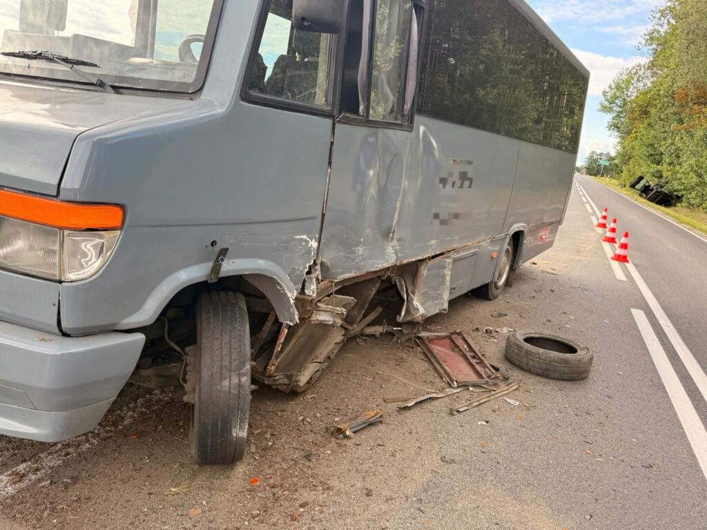 Uszkodzony autobus stojący na poboczu drogi, z odłamanym kołem i uszkodzoną karoserią, obok ustawione pachołki drogowe.