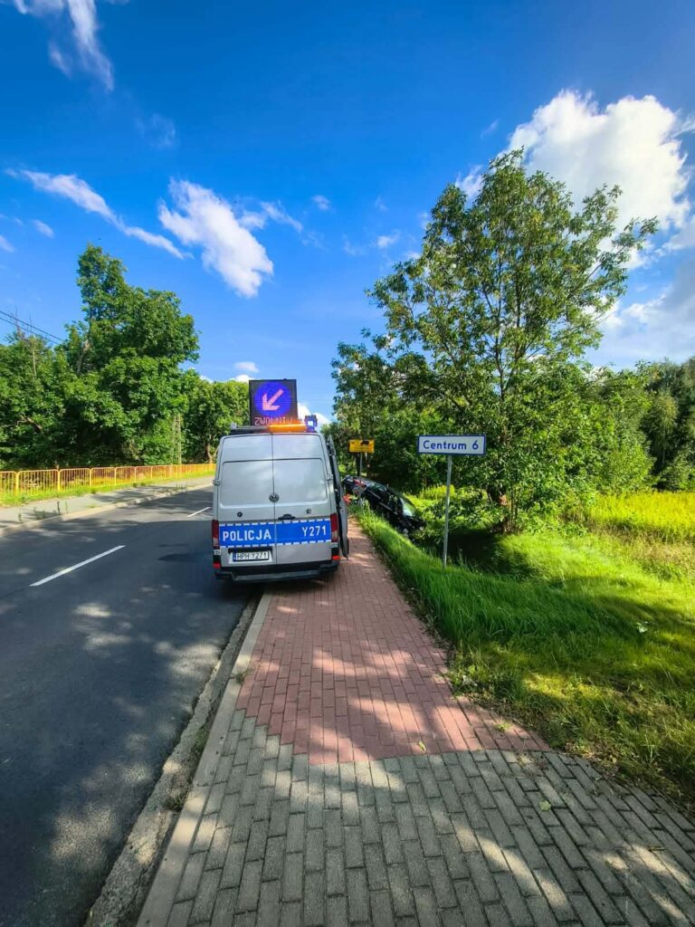Policyjny radiowóz zaparkowany na chodniku, obok znaku kierunkowym do centrum, w otoczeniu zieleni.