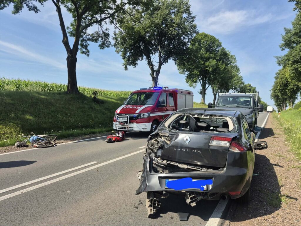 Na drodze leży uszkodzony rower, obok niego leży osoba. W tle widoczny jest czerwony wóz strażacki oraz rozbity samochód osobowy.