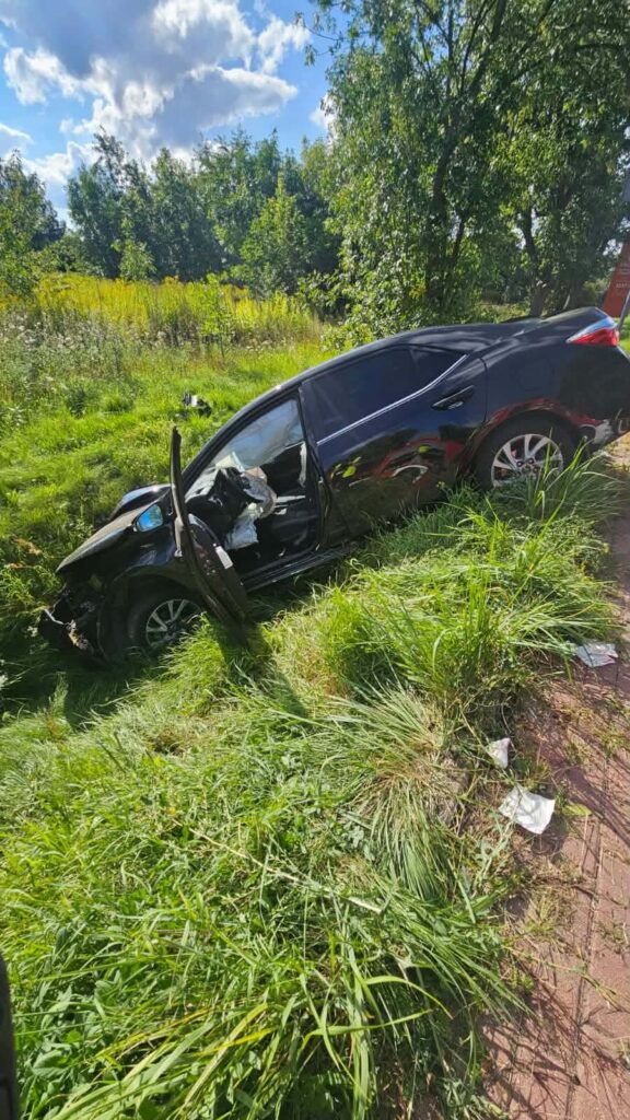 Czarny samochód osobowy w rowie, z otwartymi drzwiami, otoczony wysoką trawą i zaroślami.