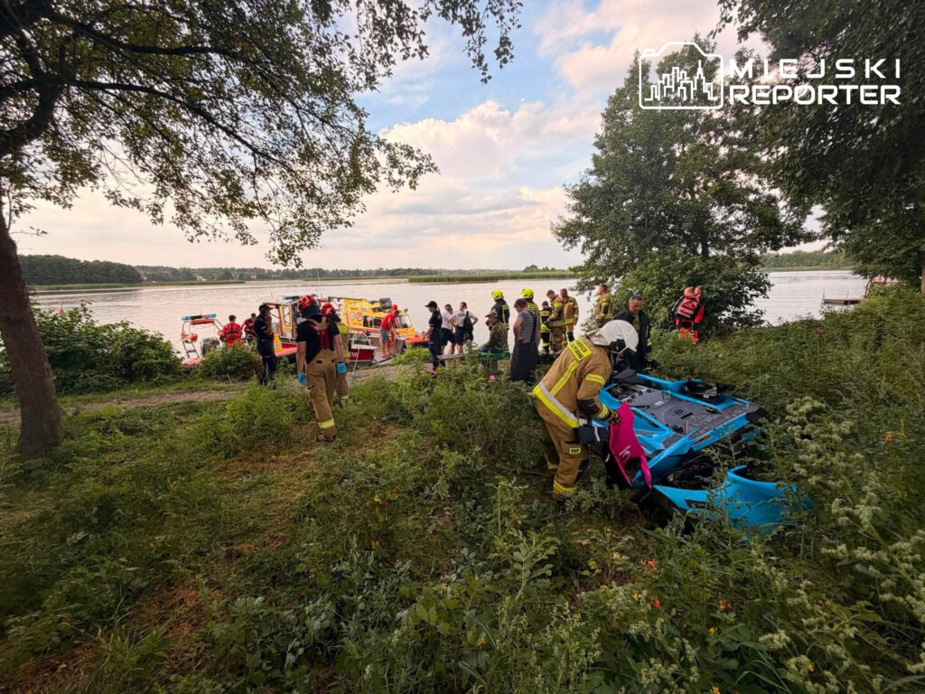 Grupa ratowników w mundurach pracuje nad przewróconym pojazdem w pobliżu wody, otoczona zielenią.
