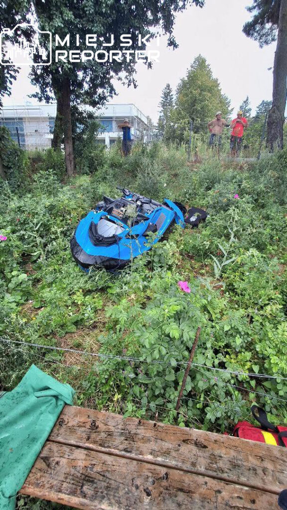 Niebieska motorówka leży przewrócona w gęstej zieleni, a w tle widać grupę trzech mężczyzn obserwujących sytuację.