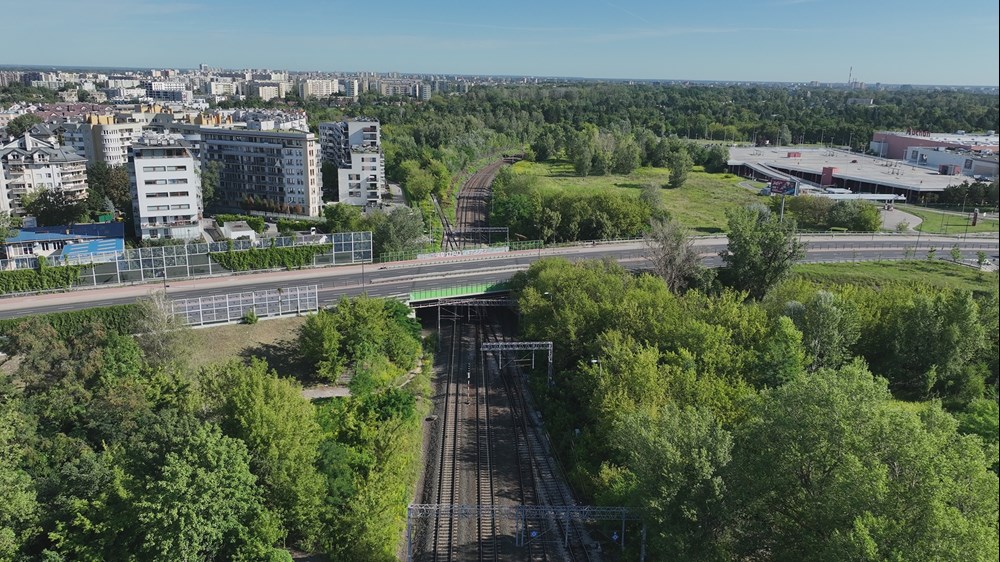 Widok na tory kolejowe otoczone zielenią, w tle nowoczesne budynki mieszkalne i centrum handlowe.
