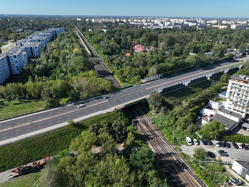 Widok z lotu ptaka na dwupasmową drogę, obok torów kolejowych, otoczoną zielenią i nowoczesnymi budynkami mieszkalnymi.