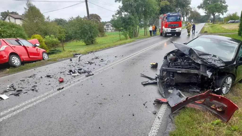 Na drodze leży rozbity czarny samochód osobowy z uszkodzonym przodem, obok czerwony pojazd na poboczu; wokół fragmenty karoserii.