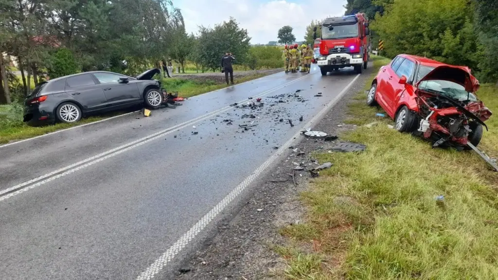 Na drodze leżą resztki zderzenia dwóch samochodów, w tym czarnego hatchbacka i czerwonego pojazdu z uszkodzoną przednią częścią. W tle widać strażaków i radiowóz.