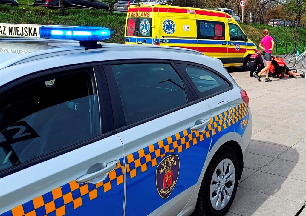 Strażnicy miejscy udzielają pomocy mężczyźnie leżącemu na chodniku obok radiowozu i karetki.