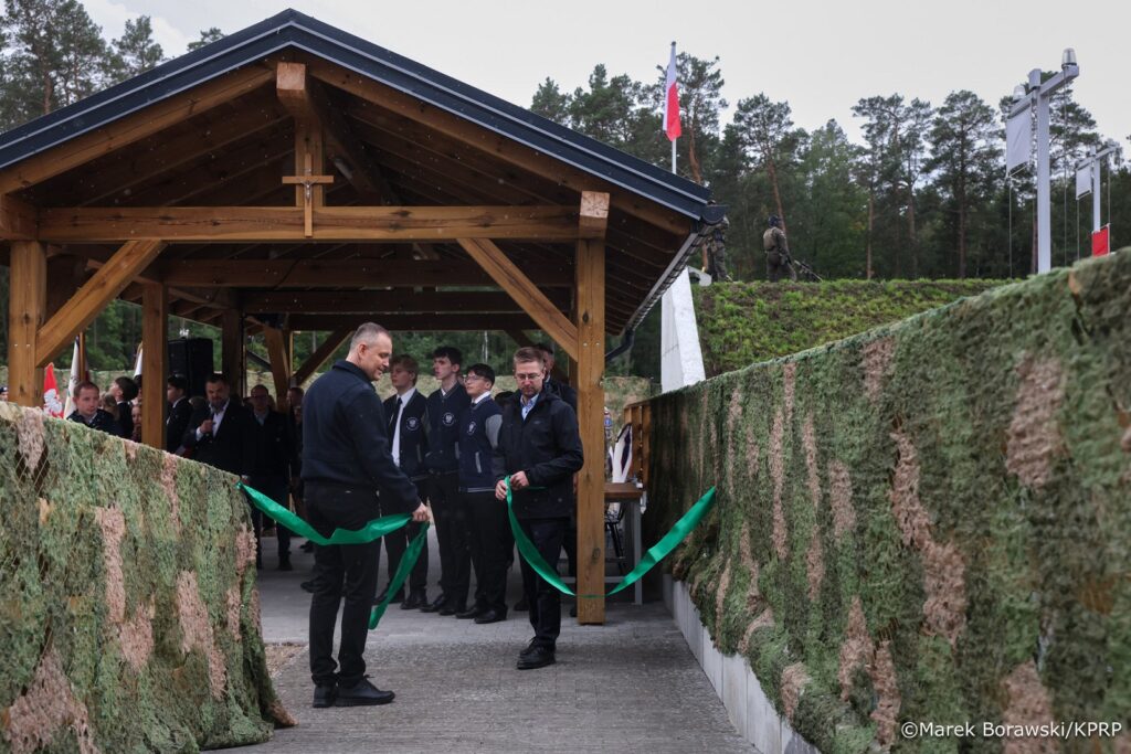 Mężczyzna w czarnej odzieży przerywa zieloną wstęgę podczas ceremonii otwarcia w drewnianym pawilonie w lesie.