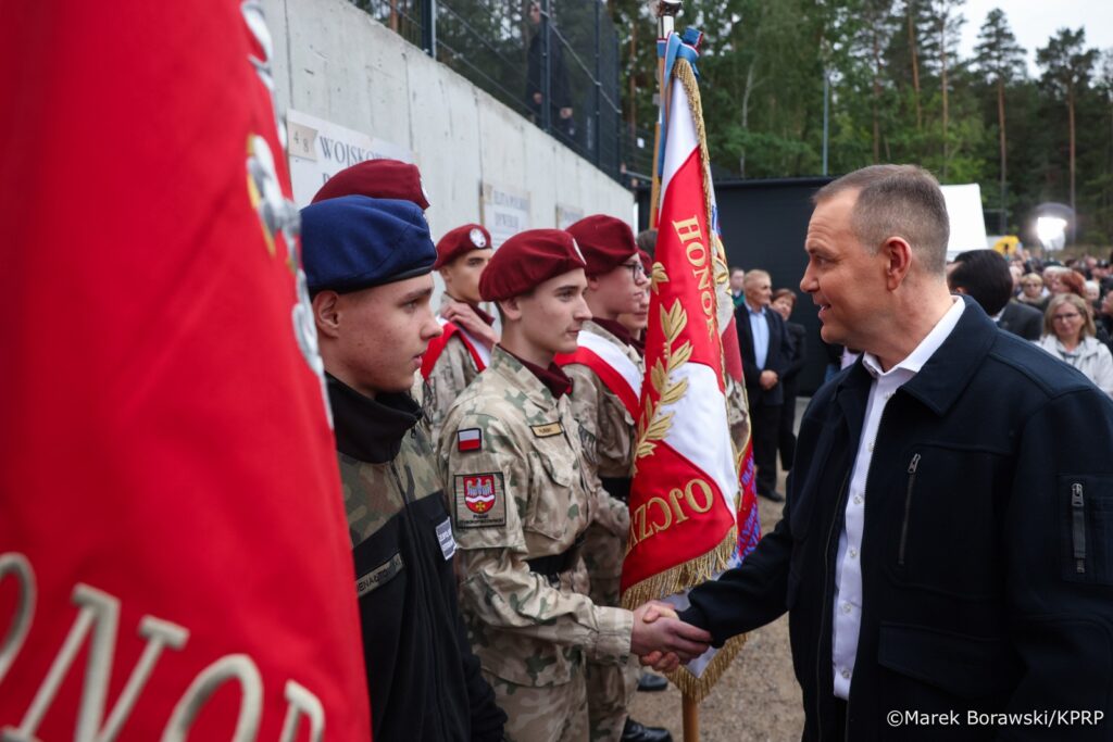 Mężczyzna w ciemnej kurtce uścisnął dłoń żołnierza w mundurze, stojąc obok flagi w kolorze czerwonym z białymi detalami.