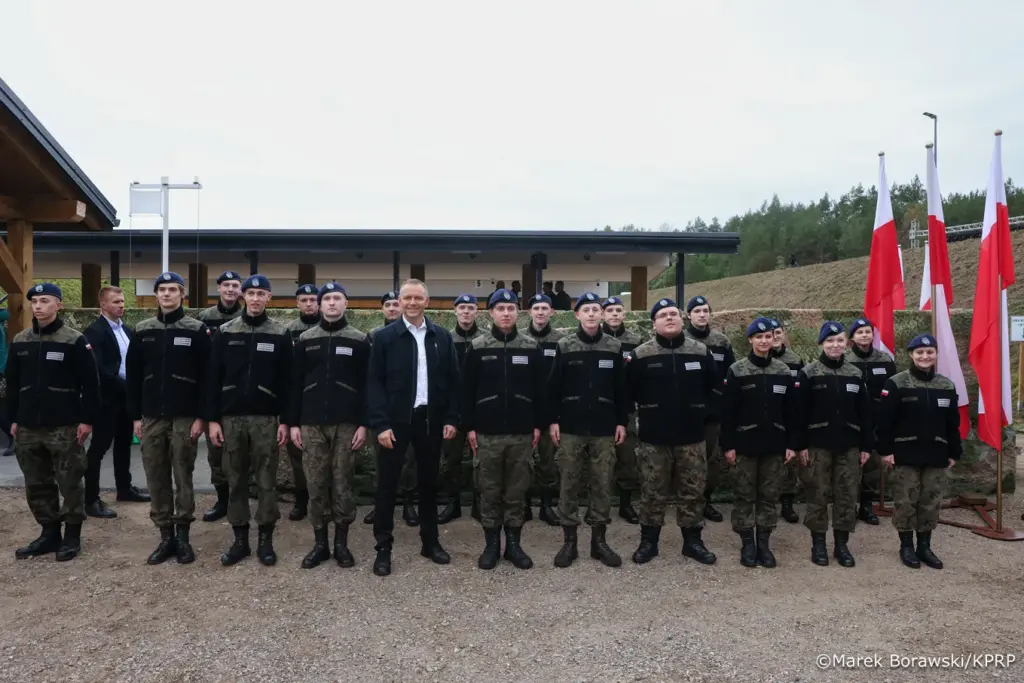 Grupa żołnierzy w mundurach i beretach stoi w szeregu przed nowoczesnym budynkiem, w tle flagi narodowe.