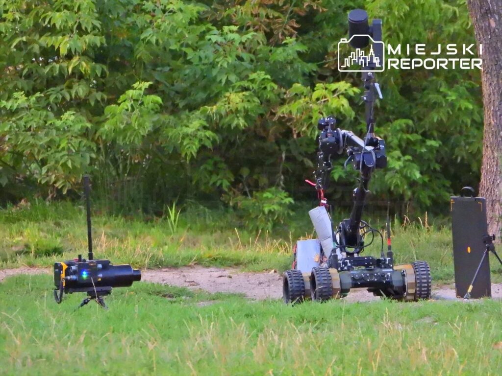 Robot zdalnie sterowany z kamerą i sprzętem technicznym na trawie, w otoczeniu zielonych krzewów.
