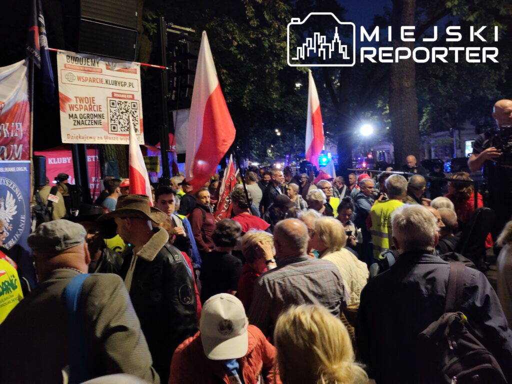 Tłum ludzi z flagami narodowymi i transparentami zbiera się na ulicy w nocy, otoczeni przez drzewa i oświetlenie.