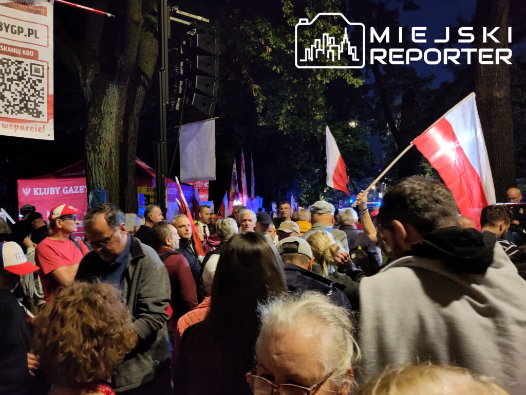 Tłum ludzi z flagami Polski gromadzi się w ciemnym otoczeniu, obok stoisk z transparentami.