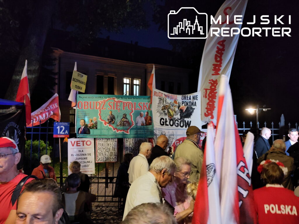 Grupa ludzi z flagami i transparentami protestuje w nocy przed budynkiem, na którym widnieje hasło "OBUDŹ SIĘ POLSKO".