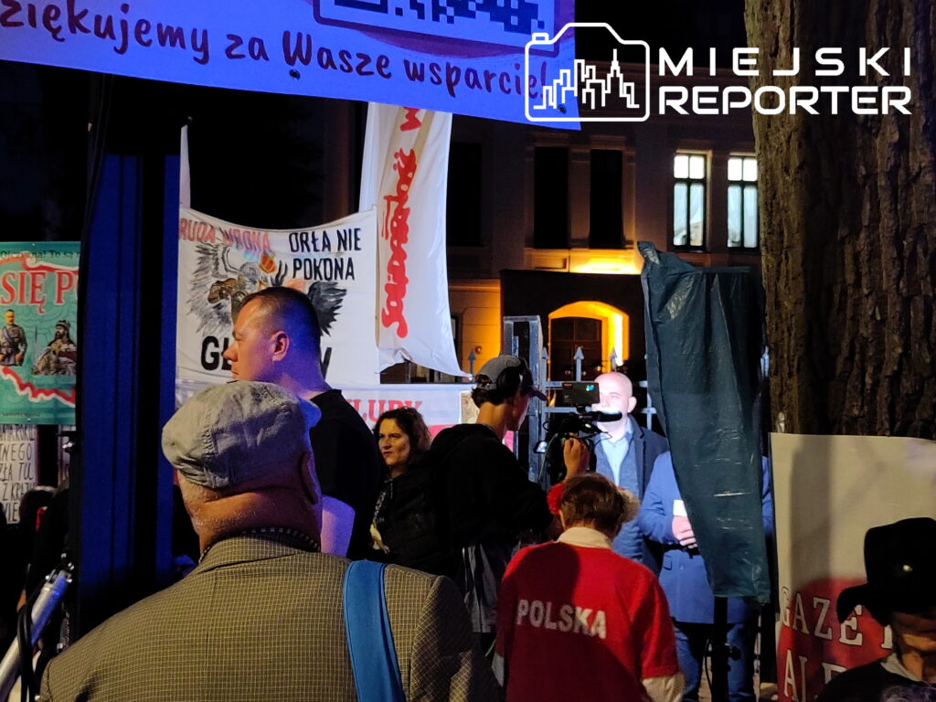 Grupa ludzi w tle z transparentami, mężczyzna z kamerą nagrywający rozmowę przy oświetlonym budynku.