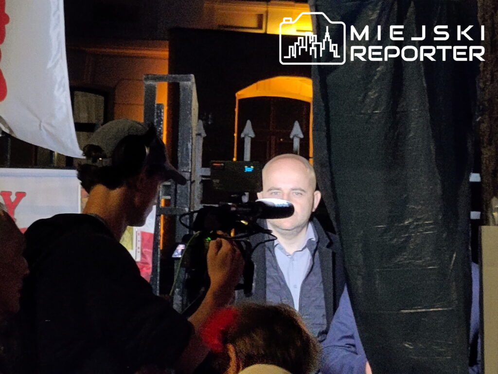 Mężczyzna z mikrofonem stoi na scenie, podczas gdy operator kamery rejestruje wydarzenie w tle.