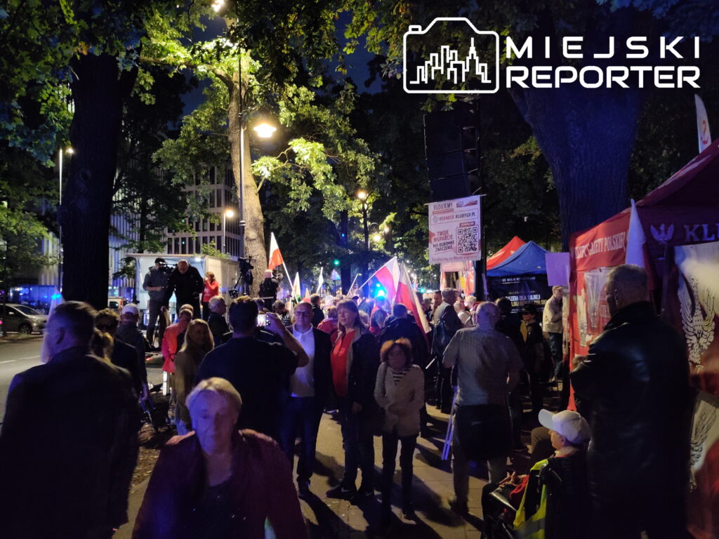 Tłum ludzi z flagami narodowymi i transparentami, zorganizowani na nocnym wydarzeniu w miejskim otoczeniu.