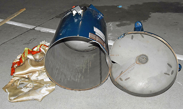 Zniszczony pojemnik metalowy leży na ziemi obok fragmentów materiału. W tle widać betonową nawierzchnię.