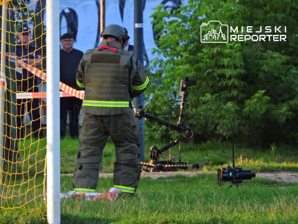 Mężczyzna w mundurze ochronnym obsługujący sprzęt saperski na trawie, z widokiem na grupę obserwatorów w tle.