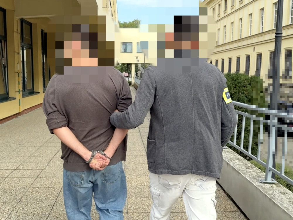 Mężczyzna w kajdankach prowadzony przez funkcjonariusza policji na chodniku w miejskim otoczeniu.