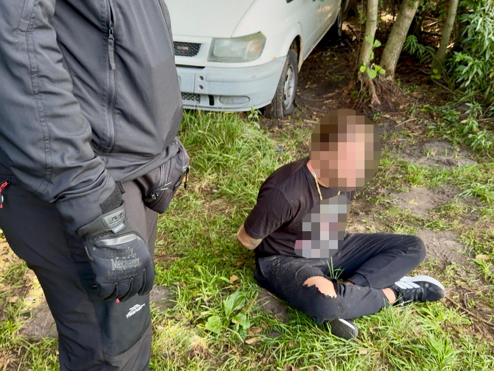 Funkcjonariusz w czarnym stroju stoi obok młodego mężczyzny w czarnej koszulce, siedzącego na trawie z rękami związanymi z tyłu.