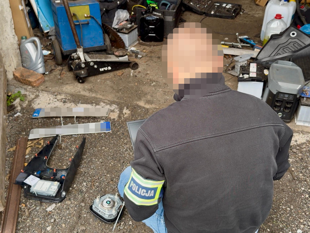 Funkcjonariusz policji przegląda laptopa, siedząc na ziemi w zagraconym warsztacie, otoczony narzędziami i częściami.