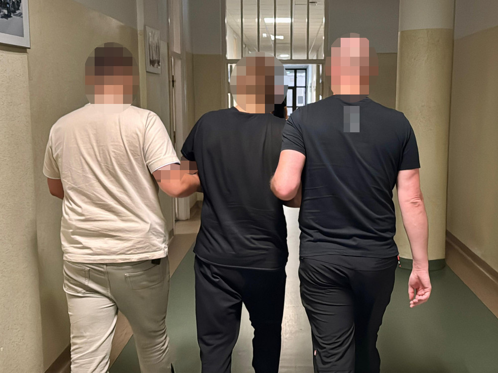Trzy osoby idą korytarzem, trzymając się za ręce, w tle widoczne drzwi i ściany z jasnym wykończeniem.