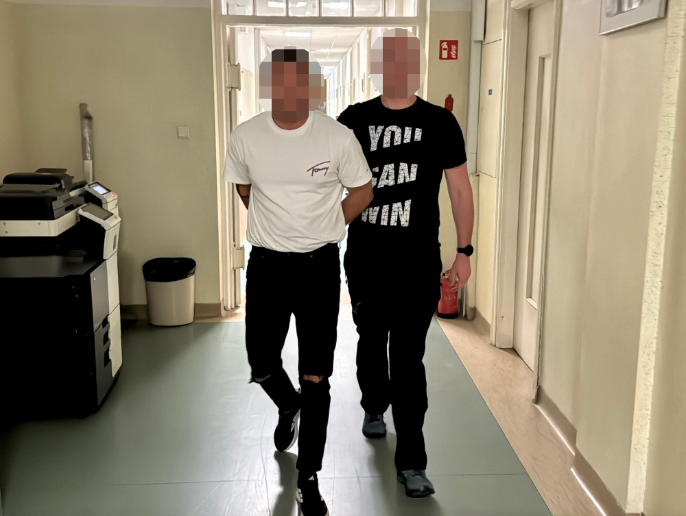 Mężczyzna w białej koszulce prowadzony przez drugiego mężczyznę w czarnej koszulce w korytarzu budynku.