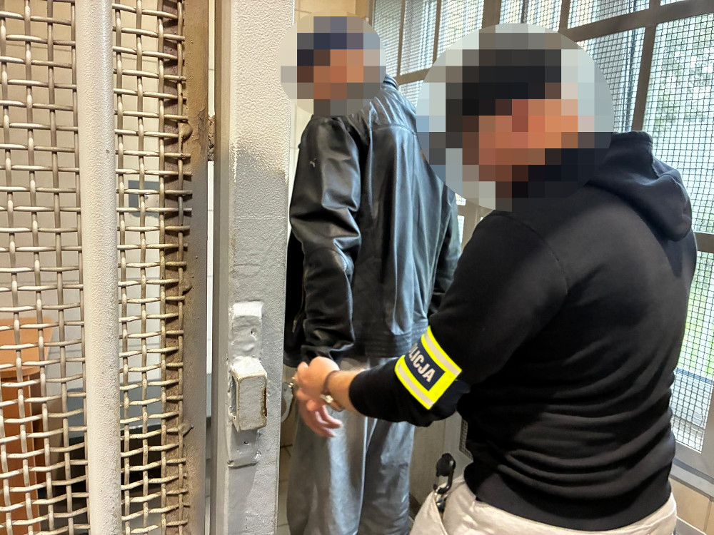 Mężczyzna w skórzanej kurtce jest zatrzymywany przez funkcjonariusza policji w pomieszczeniu z metalowymi kratami.