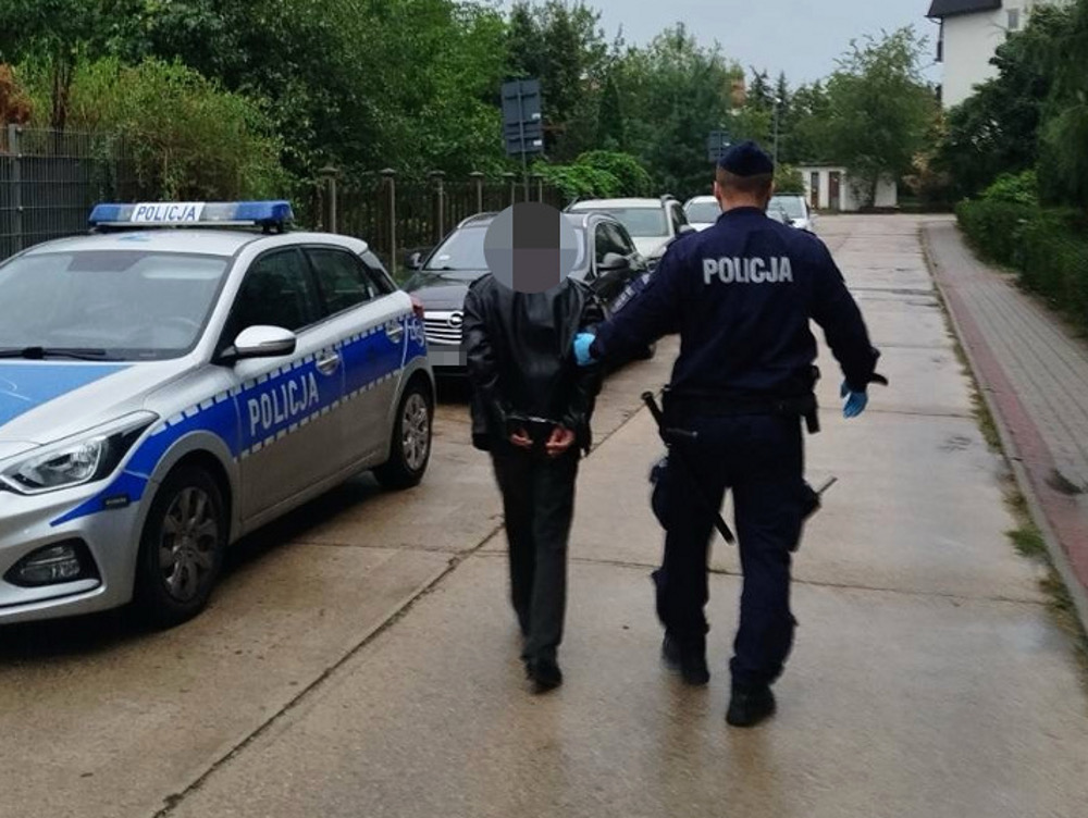 Mężczyzna w rękach policjanta prowadzony wzdłuż ulicy, obok radiowozu policyjnego.