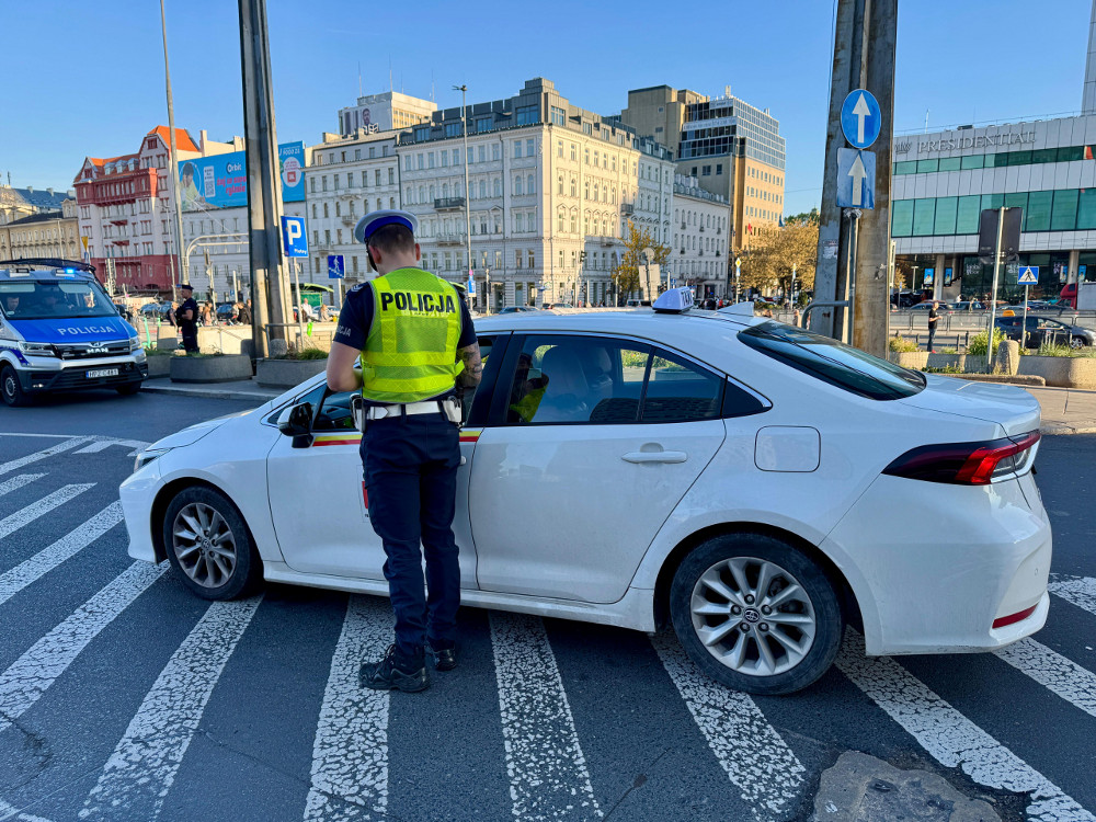 Policjant w kamizelce z napisem „POLICJA” kontroluje kierowcę białego samochodu na skrzyżowaniu wśród miejskiej zabudowy.