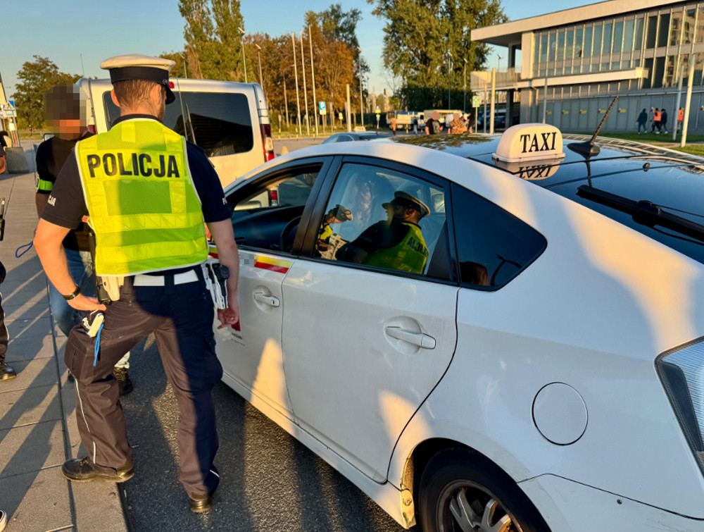 Policjant w kamizelce z napisem „POLICJA” kontroluje kierowcę białej taksówki przy ulicy.