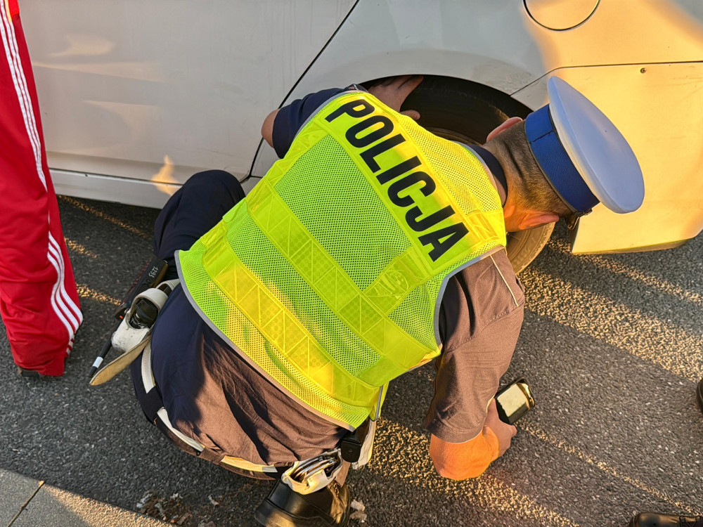 Policjant w odblaskowej kamizelce z napisem „POLICJA” pochyla się przy otwartym samochodzie, obok stoi osoba w czerwonym
