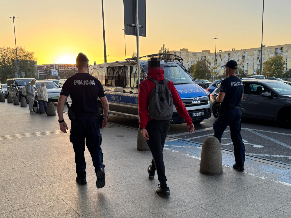 Dwóch policjantów prowadzi osobę w czerwonej bluzie w stronę oznakowanego radiowozu na parkingu o zachodzie słońca.