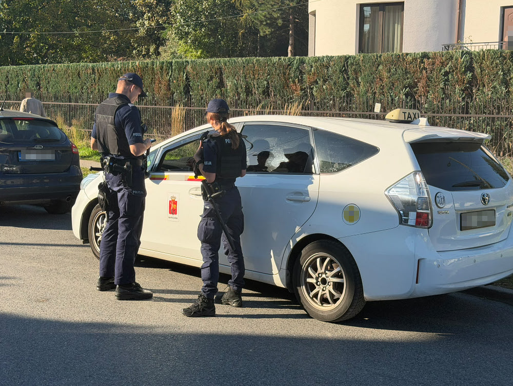 Dwóch umundurowanych policjantów rozmawia przy białej taksówce zaparkowanej przy ogrodzeniu budynku.