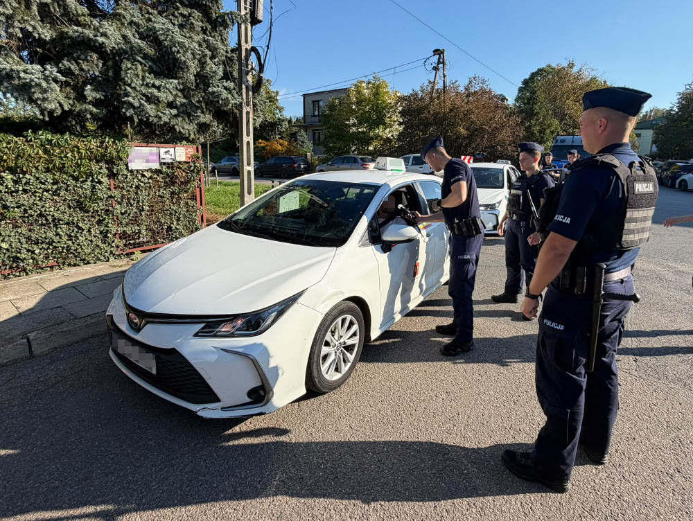 Policjant rozmawia z kierowcą przez otwarte okno białego samochodu, a obok stoją funkcjonariusze.